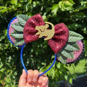 May include: A headband featuring two circular ears covered in light pink sequins with a dark blue pom-pom trim. A large, central bow made of dark red sequins is adorned with a gold glitter silhouette of a lion cub. Green fabric leaves.
