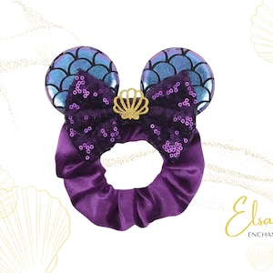 Puede incluir: Coletero morado con un lazo de lentejuelas y orejas azules iridiscentes, coronado con una corona dorada. El diseño evoca el tema de las sirenas. El texto "Elsa's Enchantments" está en la esquina inferior derecha.