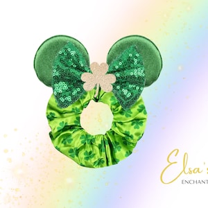 May include: A green St. Patrick's Day themed hair accessory with two round ears, a sequined bow, and a gold glitter shamrock. The scrunchie is green with a clover pattern. The text "Elsa's Enchantments" is in gold script.