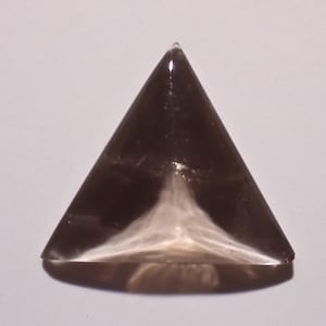 Puede incluir: Una gema triangular pulida, de color marrón oscuro, con un patrón arremolinado más claro en su base. La gema está tallada con una base plana y un ápice puntiagudo, mostrando su forma geométrica y variaciones naturales.
