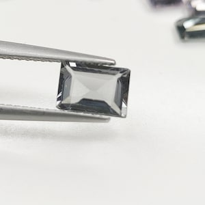 May include: A rectangular, faceted, translucent grey gemstone held by metal tweezers. The stone features a clear, geometric cut with sharp edges and a polished surface, reflecting light across its multiple angled facets in a precise, symmetrical pattern.
