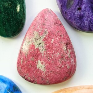 Peut inclure: Une pierre précieuse cabochon rose et blanche avec une surface rugueuse et texturée. La pierre est en forme de larme et a un aspect naturel et organique.