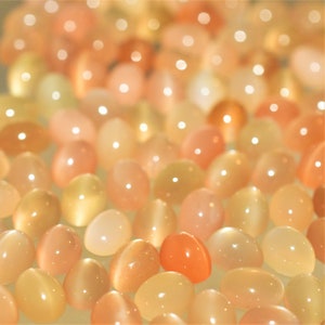 May include: A collection of polished, oval-shaped gemstones in shades of peach, orange, and cream. The stones have a smooth, glossy surface and are arranged closely together, creating a warm, inviting display. The gemstones are likely used for jewelry or decorative purposes.