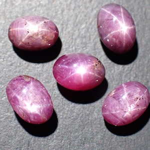 Pode incluir: Cinco pedras preciosas de rubi estrela ovais. As gemas são de uma cor vermelho-rosada profunda, com um efeito de estrela de seis raios visível na superfície. Cada pedra tem uma aparência polida e lisa, com inclusões naturais.