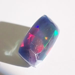 May include: A faceted, rectangular black opal gemstone. The stone displays flashes of red, green, and blue iridescence. The gem is set against a white background, with a shadow cast to the left.