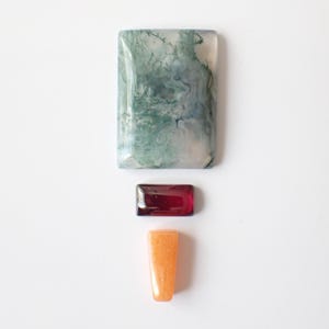 May include: A rectangular moss agate cabochon with green dendritic inclusions, a smaller rectangular deep red garnet cabochon, and a small trapezoidal orange aventurine stone arranged vertically on a plain white surface.