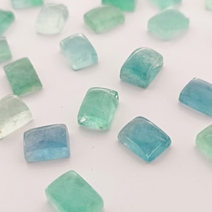May include: A scattering of rectangular, polished gemstones in shades of blue and green. The stones have rounded edges and a translucent appearance, set against a white backdrop. The gems vary in colour from light teal to deep turquoise.