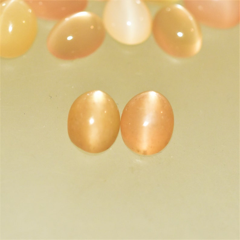 May include: A collection of oval-shaped gemstones in shades of peach and cream. The gemstones have a smooth, polished surface with a subtle cat's eye effect. The stones are arranged on a light yellow surface.