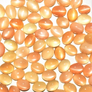 May include: A collection of oval-shaped, peach-colored gemstones with a cat's eye effect. The stones vary in shade from pale yellow to deep orange, with a glossy, polished finish. These gemstones are ideal for jewelry making.