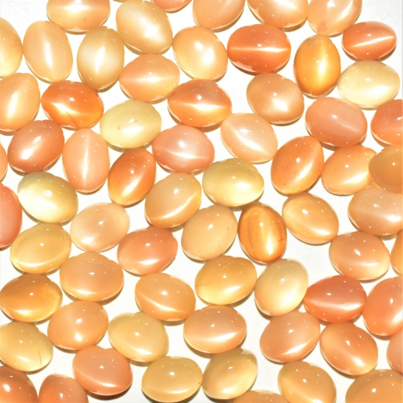 May include: A collection of oval-shaped, peach-colored gemstones with a cat's eye effect. The stones vary in shade from pale yellow to deep orange, with a glossy, polished finish. These gemstones are ideal for jewelry making.
