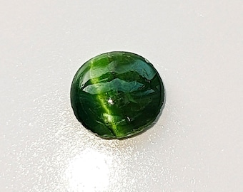 Puede incluir: Una gema redonda y pulida de color verde oscuro. La gema tiene una superficie lisa y reflectante con patrones internos visibles. La piedra está sobre un fondo blanco, resaltando su color y forma. Esta gema podría usarse en joyería.