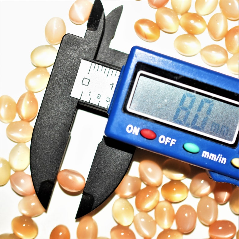 May include: A collection of oval-shaped, peach-colored gemstones. A blue digital caliper displays a measurement of 8.0 mm. The gemstones are scattered on a white surface, with varying shades of peach and orange.