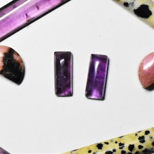 Puede incluir: Dos piedras preciosas rectangulares de amatista pulida con un tono púrpura intenso están centradas en un fondo blanco. Varias otras piedras preciosas pulidas en tonos de rosa, púrpura y amarillo con manchas negras rodean las piedras de amatista.