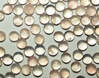May include: A collection of round, translucent, light pink and white gemstone cabochons. The cabochons are scattered on a light gray surface.