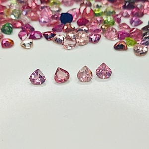 May include: Four pink, heart-shaped gemstones with a slight purple hue. The gemstones are arranged in a row on a white surface.