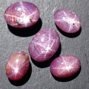 Könnte beinhalten: Fünf ovale Sternrubin-Edelsteine. Die Edelsteine haben eine tiefrot-violette Farbe mit einem sechsstrahligen Sternmuster auf der Oberfläche. Einige Steine weisen kleine Einschlüsse und Farbvariationen auf.