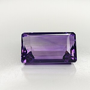 Puede incluir: Una gema de amatista tallada en forma rectangular, de color púrpura intenso. Las facetas de la piedra reflejan la luz, creando un efecto brillante. La gema se encuentra sobre un fondo blanco, resaltando su color y claridad. Para joyería.