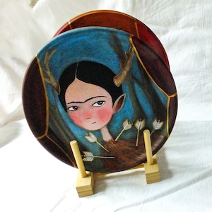 Puede incluir: Un plato de cerámica pintado a mano con un diseño azul, rojo y dorado. El plato presenta un retrato de una mujer con una diadema de asta de ciervo y flechas que atraviesan su cuerpo. El plato se exhibe en un soporte de madera.