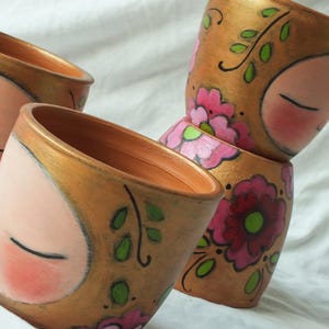 Puede incluir: Una pila de cuatro macetas de cerámica pintadas a mano. Cada maceta presenta una cara con los ojos cerrados, mejillas rosadas y un borde dorado. Las macetas están decoradas con flores rosas y hojas verdes sobre un fondo dorado.