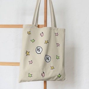 Heartstopper Leaves Tote Bag in natural cotton hanging on a hook. Heartstopper leaves printed on both sides of the bag along with two Hi quotes from the show.