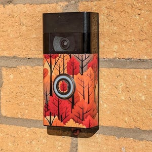 May include: A black doorbell camera with a colourful autumn leaves design skin. The camera has a round lens and a white button.