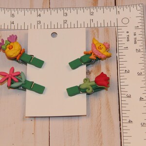 May include: Four decorative green clips with colorful floral and bird designs. The clips are attached to a white card, with a ruler in inches and a wooden background.
