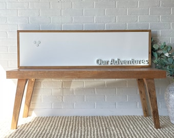 May include: A white framed whiteboard with the words "Our Adventures" written in white letters. The whiteboard is sitting on a wooden bench with a light brown rug underneath.