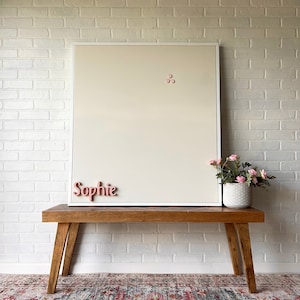 Puede incluir: Un tablero magn&eacute;tico blanco enmarcado con el nombre "Sophie" escrito en letras rosas. El tablero est&aacute; montado en un banco de madera con una planta en maceta delante.