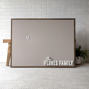 May include: A large, rectangular, framed bulletin board with a neutral-colored surface. The wooden frame surrounds the board. The words "the FLORES FAMILY" are displayed in white, three-dimensional letters. Three white magnets are attached to the board.