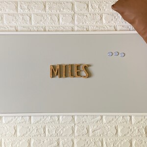 Puede incluir: Un tablero de corcho enmarcado en blanco con el nombre "MILES" escrito en letras de madera. Tres chinchetas azules est&aacute;n en el tablero.