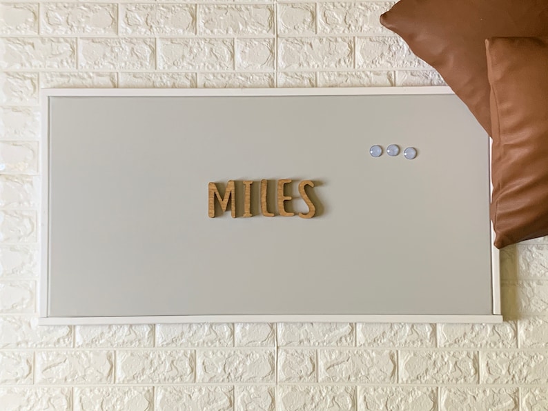 Puede incluir: Un tablero de corcho enmarcado en blanco con el nombre "MILES" escrito en letras de madera. Tres chinchetas azules est&aacute;n en el tablero.