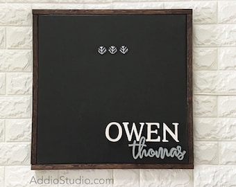 Puede incluir: Una pizarra enmarcada en marrón oscuro con el nombre "OWEN thomas" escrito en letras blancas y grises. Hay tres pequeños detalles decorativos plateados sobre el nombre.