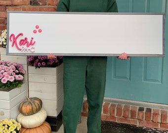 May include: A person holding a white sign with the text "Kari PA-C" in pink letters. The sign is framed in gray and has a few pink dots on the top left corner.