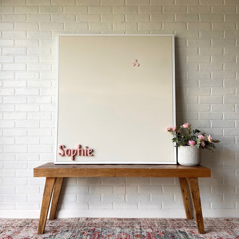 Puede incluir: Un tablero magn&eacute;tico blanco enmarcado con el nombre "Sophie" escrito en letras rosas. El tablero est&aacute; montado en un banco de madera con una planta en maceta delante.