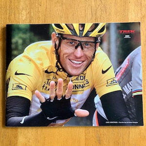 May include: A close-up of a cyclist wearing a yellow jersey and a helmet, smiling and waving. The cyclist is wearing a black glove and a white arm band. The text "TREK" and "2004" are visible in the top right corner of the image. The text "LANCE ARMSTRONG - Five-time Tour de France champion" is visible at the bottom of the image.