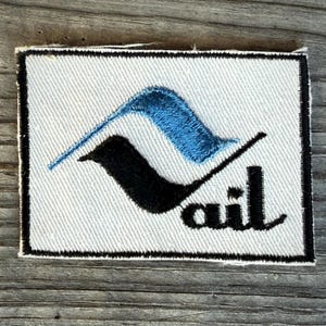May include: A rectangular patch with a white background and a black border. The patch features a blue and black stylized design and the word "vail" in black script.