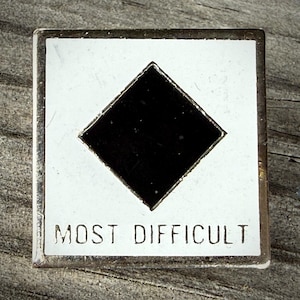 May include: A square-shaped metal pin with a white enamel background and a centered black diamond shape. The text MOST DIFFICULT is printed in black capital letters along the bottom edge of the pin, which features a metallic silver-toned border.