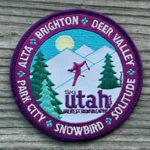May include: A circular embroidered patch with a purple border featuring the text "Alta, Park City, Brighton, Deer Valley, Solitude, Snowbird". The patch has a skier, mountains, trees, and the words "Ski Utah".