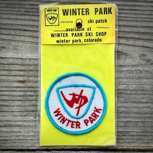 May include: A vintage Winter Park ski patch on a yellow backing. The patch is circular with a white background, a blue border, and red and blue text. The text on the patch reads "Winter Park". The backing has text that reads "Winter Park Ski Shop".
