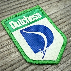 May include: Embroidered patch with the word "Dutchess" in white on a green background. The patch features a blue and white design with a skier. The patch has a green border and is on a wooden surface.