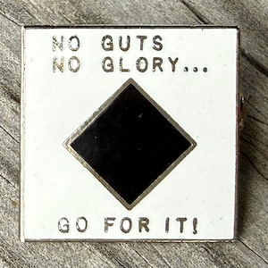 May include: A square metal enamel pin featuring a white background with a centered black diamond shape. The text NO GUTS NO GLORY is printed in black above the diamond, and the text GO FOR IT! is printed in black below the diamond shape.