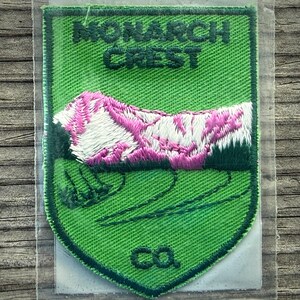 May include: Green embroidered patch with the words "MONARCH CREST" and a design of a pink and white mountain range. The patch has a shield shape and the letters "ca" at the bottom.