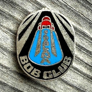 May include: Oval-shaped Bob Club pin with a black and silver design. The pin features a blue central design with the text "SIX PLAZA" in silver. The top of the design is red. The words "BOB CLUB" are in silver around the bottom.