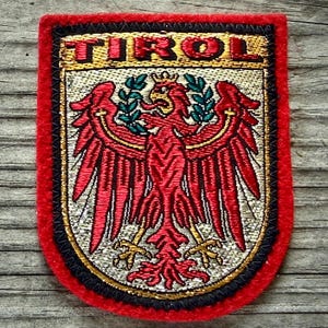 May include: Embroidered patch featuring the word "TIROL" above a red eagle emblem with outstretched wings, holding a green sprig. The patch has a red border with black and gold accents, and a silver background.