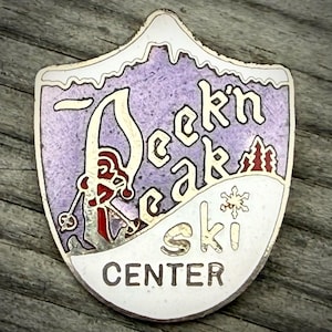 May include: A vintage ski center pin in a shield shape. The pin features the words "Deek'n Peak Ski Center" in a stylized font, with a skier graphic and a snowflake. The background is purple and white, with a silver border.