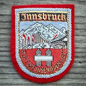 May include: Embroidered patch with the word "Innsbruck" at the top. The patch features a mountain scene with ski lifts and the text "RUFT DIE JUGEND DER WELT" in gold lettering on a red background, framed by a red border.