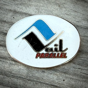 May include: Oval-shaped pin with a white background and a silver border. The pin features a stylized design in blue and black, with the word "ail" in black and "PARALLEL" in red.