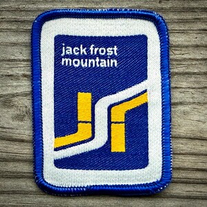May include: Embroidered patch with a blue border, white background, and blue center. The text "jack frost mountain" is in white. The design includes yellow and white geometric shapes. The patch is rectangular with rounded corners.
