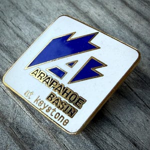 May include: A square-shaped enamel pin with a white background and a gold-toned metal border. The pin features a blue geometric mountain graphic above the gold-colored text that reads ARAPAHOE BASIN at keystone in a bold, sans-serif font.