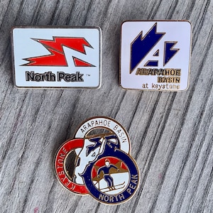 May include: Three vintage enamel pins featuring the Arapahoe Basin ski resort in Colorado. The first pin is white with a red and white logo that says "North Peak". The second pin is white with a blue logo that says "Arapahoe Basin at Keystone". The third pin is a circular pin with a red, white, and blue logo that says "Arapahoe Basin North Peak Keystone".